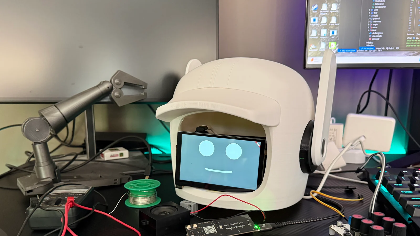 Droid head prototype on a workbench with tools, wiring, and code visible on a monitor